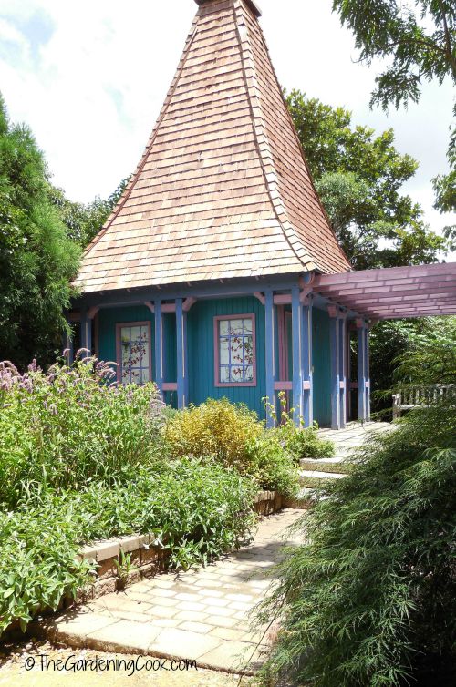 Pequena casa azul com telhado curvo, cercada por bordas cheias e um caminho.