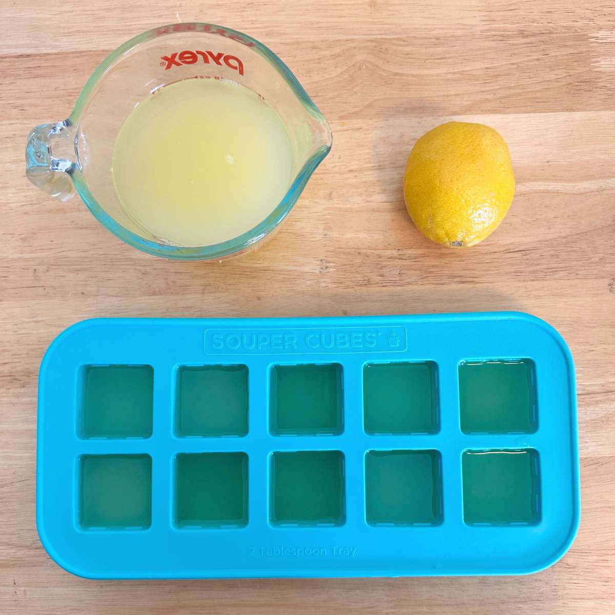 Uma imagem de processo de congelar suco de limão em formas de gelo de silicone que mostra um recipiente Pyrex de uma xícara cheio de suco de limão ao lado de um limão, acima de uma forma de gelo azul reutilizável cheia de suco de limão.