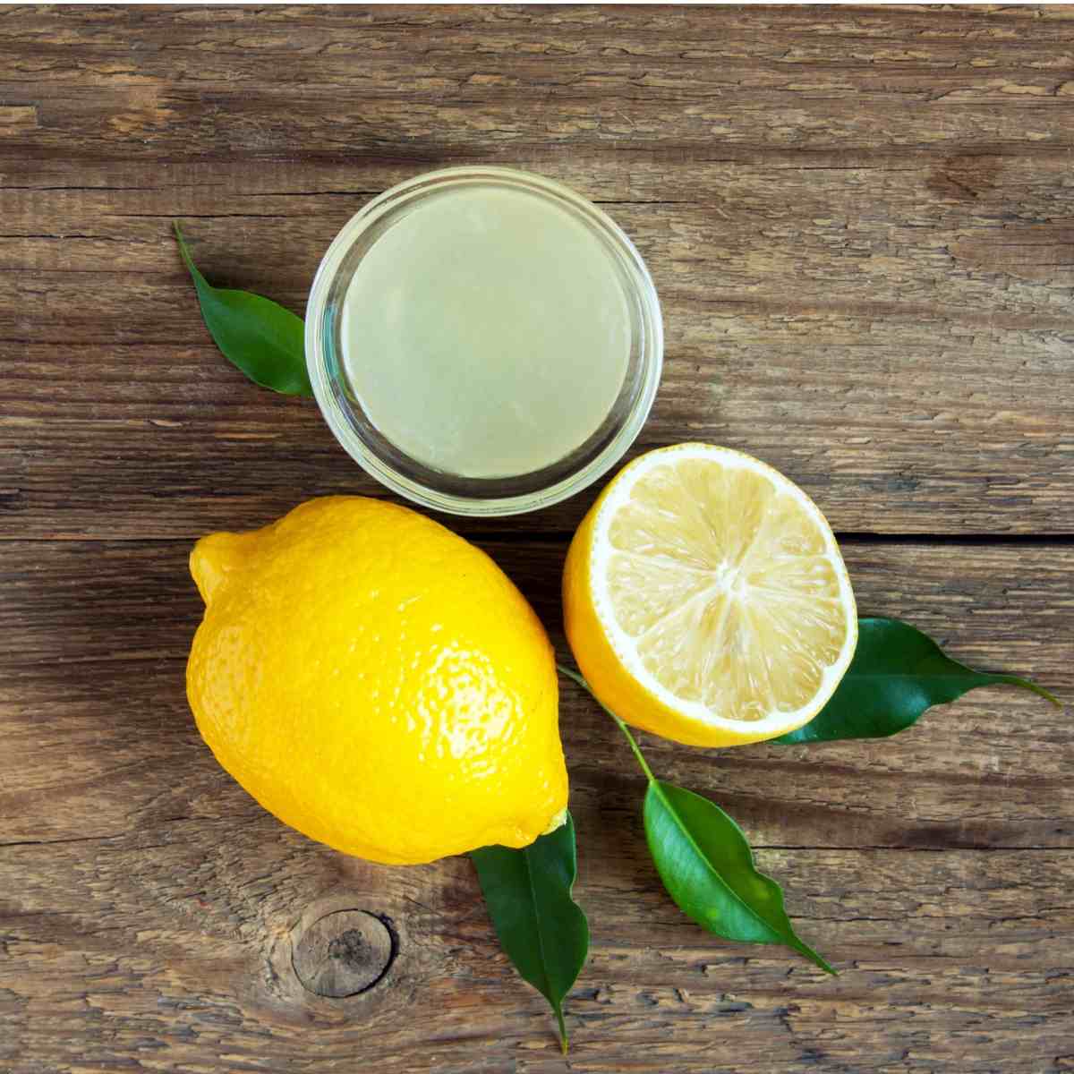 Um ramequim de vidro com suco de limão recém espremido acima de um limão inteiro e um limão cortado ao meio com várias folhas de limão ao redor.