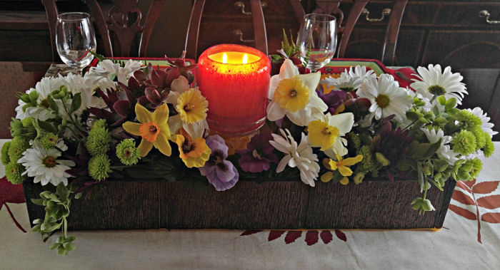 Uma caixa de madeira marrom cheia de flores de seda e uma vela acesa em um recipiente de vidro faz um lindo centro de mesa.