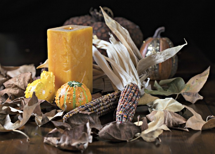 Exposição de milho indiano para uma mesa usando uma vela em bloco e abóboras em tom amarelo e espigas e folhas de milho marrons contrastantes.