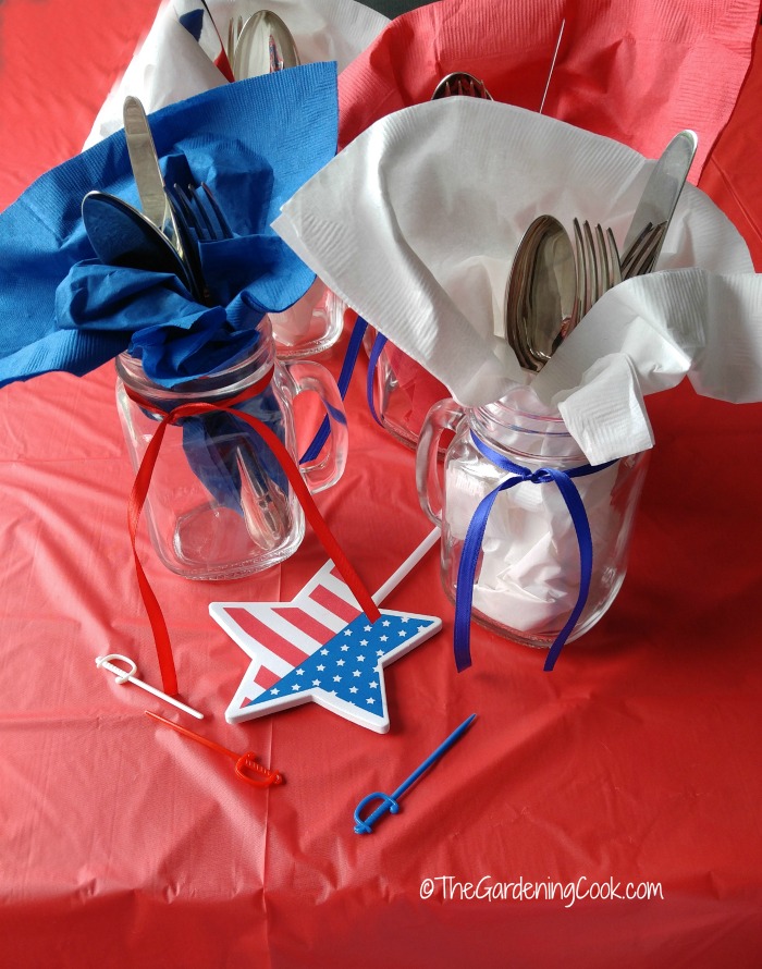 Suportes de talheres Patiotic Mason Jar com toalhas azuis e brancas, fitas vermelhas e azuis e estrelas de bandeira sobre uma toalha de mesa vermelha.