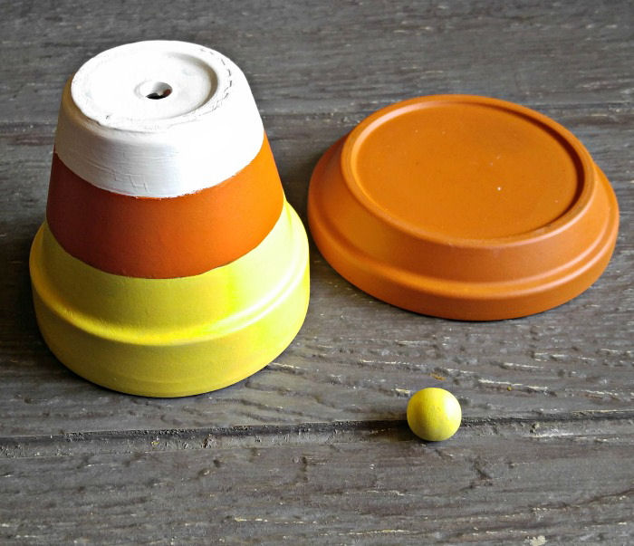 Vaso de barro pintado de laranja, amarelo e branco, saucer de barro pintado de laranja e uma pequena bola de madeira pintada de amarelo em cima de uma mesa de madeira.