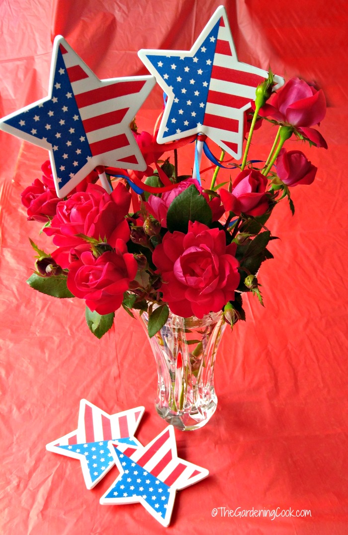 Decoração de vaso de rosas patrióticas com bandeiras estrela em uma toalha de mesa vermelha.