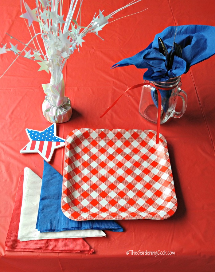 Configuração de mesa patriótica com peso de papel estrela, toalha azul em frasco de conservas, toalhas vermelhas, brancas e azuis, estrela de bandeira e pratos vermelhos e brancos.