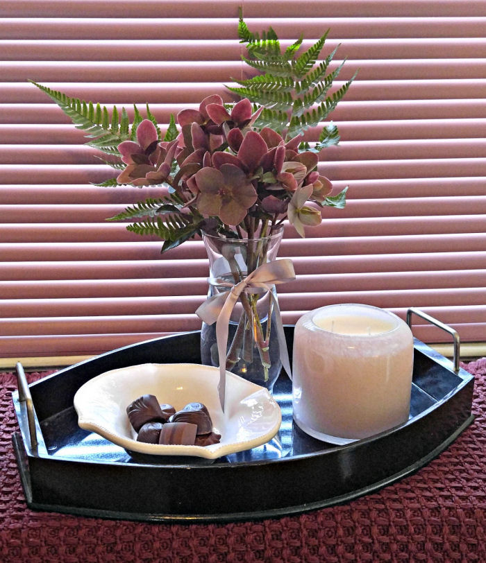 Heléboros e samambaias em um vaso de vidro, ao lado de uma tigela de chocolate e uma vela de vidro. Uma cortina rosa fechada e um caminho de mesa adicionam calor à cena.