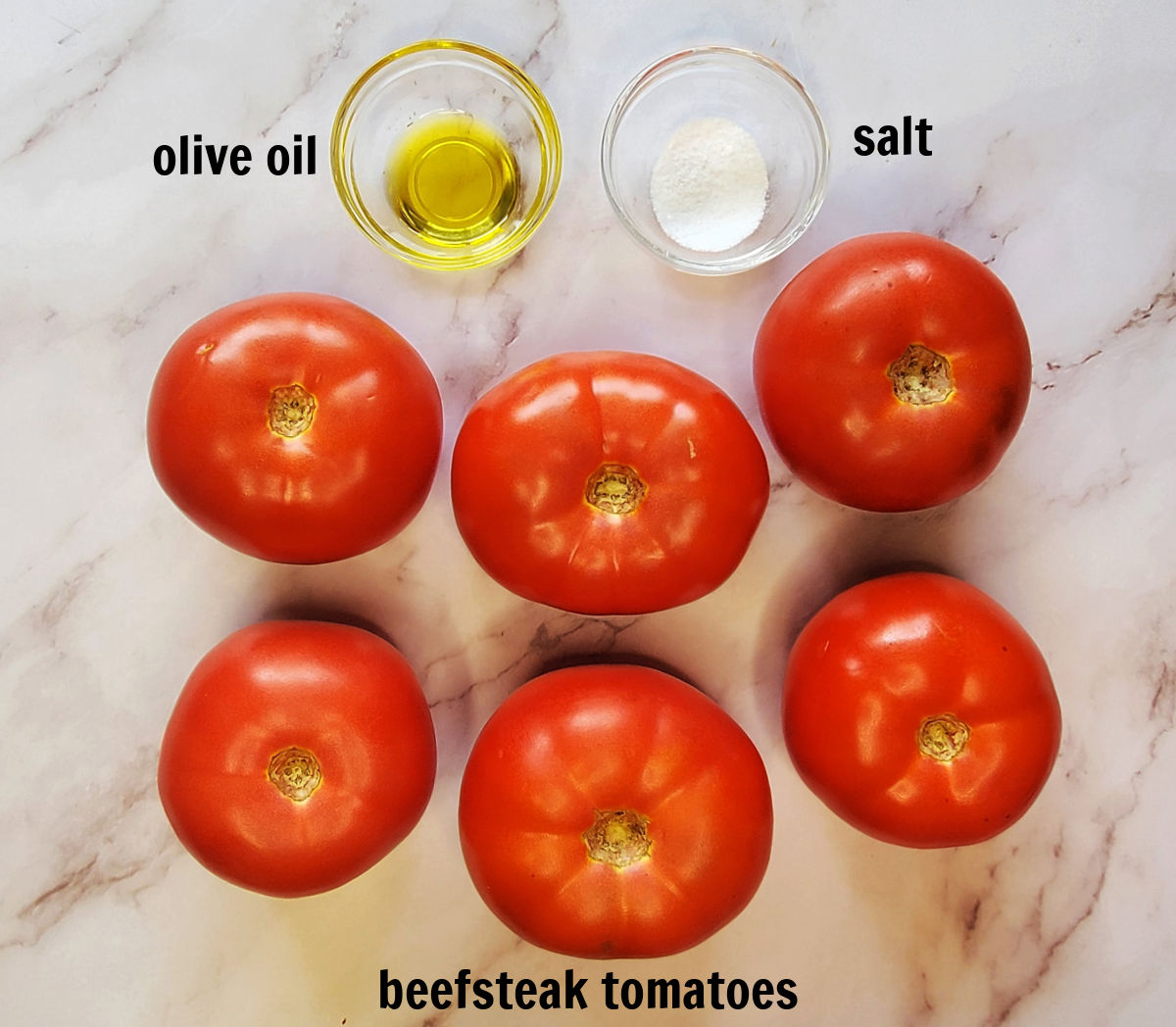 6 tomates beefsteak e duas tigelas com azeite e sal, rotuladas com texto preto, em um balcão de mármore - ingredientes para torrar tomates para molho.