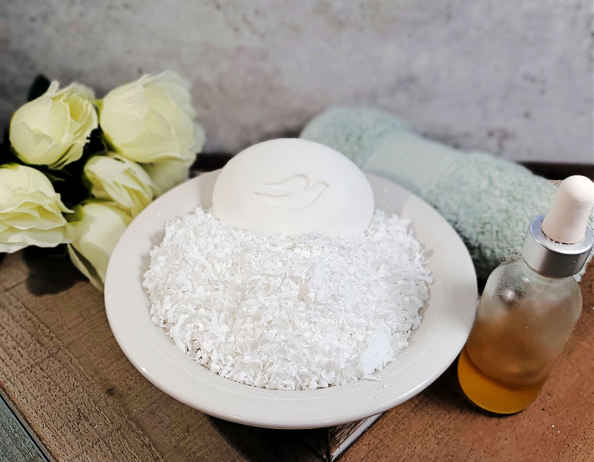 Rale as lascas de sabão Dove Uma tigela branca com lascas de sabão Dove e uma barra de sabão ao lado de rosas brancas, uma toalha verde enrolada e um frasco de óleo essencial.