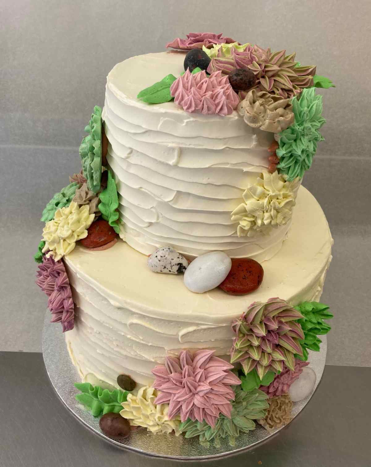 Um bolo com suculentas de buttercream com duas camadas, coberto com cobertura branca, com suculentas de buttercream (modeladas após echeveria) em rosa, amarelo, verde e marrom caindo pelas camadas do bolo.