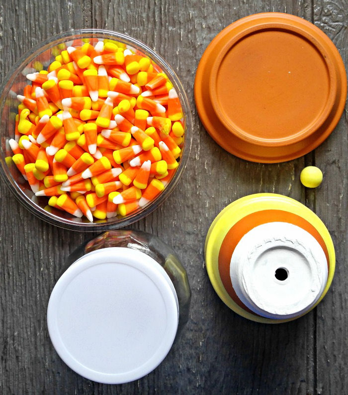 Uma tigela de vidro com doces de milho ao lado de um jarro de vidro e tampa, vaso de terracota pintado, saucer e uma pequena bola de madeira amarela.
