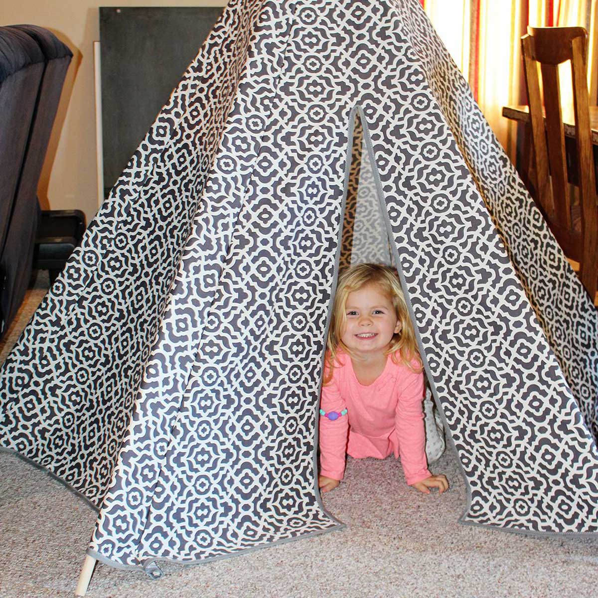 Uma criança pequena dentro de uma tenda teepee DIY feita de tecido estampado cinza e branco.